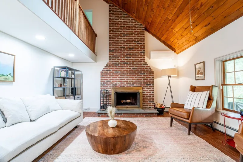 Cozy living room with a striking brick fireplace, vaulted wood ceiling, and stylish furnishings. The warm tones and comfortable seating make this space ideal for relaxing or enjoying a quiet evening by the fire.